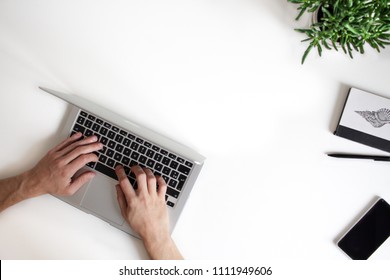 Top View Of A White Office Table, Showing Man's Hands Over A Silver Laptop Keyboard, Working On Something. Smart Phone, Pencil, Notebook  And Small Plant Are Aside. Modern Working Environment.