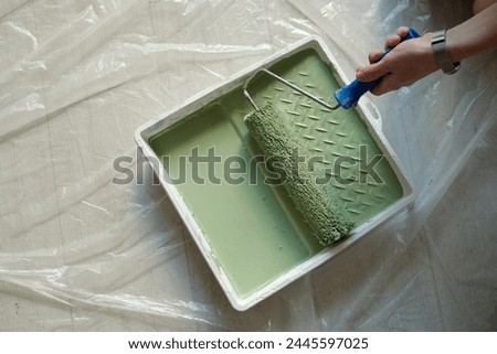 Top view of white plastic tray with green paint standing on the floor covered with cellophane and hand of woman holding paintroller