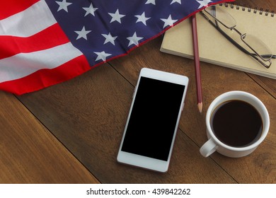 Top View The White Smartphone, Black Coffee, Pencil, Note Paper, Eyeglasses, United States Flag On Table Wood. 