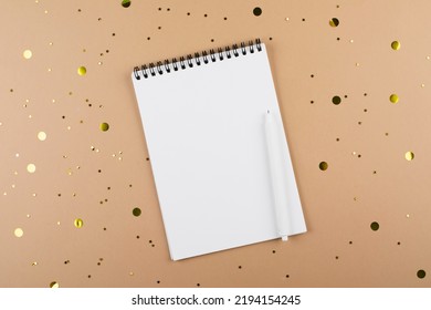 Top View Of White Spiral Notebook Note Pad And Pen On Beige Background With Golden Confetti. Mockup Concept. Copy Space.