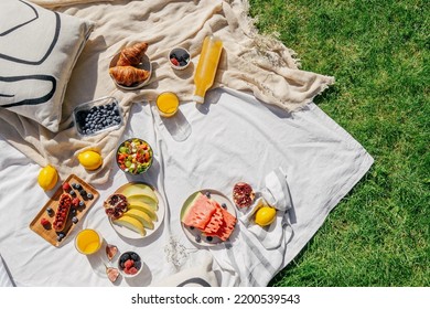 top view of white tablecloth with different food, juicy fruits, ripe berry, vegetables salad in bowl, sweet croissants, fresh orange juice in glass near bottle, plaid and pillow on grass outdoors