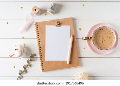 Top view of white working table background with blank paper notebook, cup of coffee. Flat lay green leaf, flowers, golden paper binder clips. Notepad and pen. Desktop mockup, workspace, stationery.