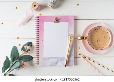 Top view of white working table background with blank paper notebook, cup of coffee. Flat lay green leaf, flowers, golden paper binder clips. Notepad and pen. Desktop mockup, workspace, stationery.