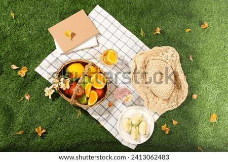 Top view of wicker picnic basket with fruits, hat and books on white checkered tablecloth on green grass outside in summer park. Advertising photo, summer day concept