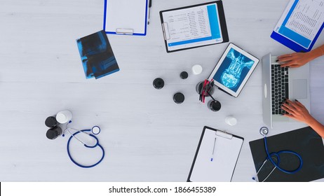 Top View Of Woman Doctor Hands Typing On Laptop Sitting On Desk With X-ray And Medical Equipment All Around. Assistant Working In Health Clinic Using Modern Devices. Copy Space, Flat Lay Concept