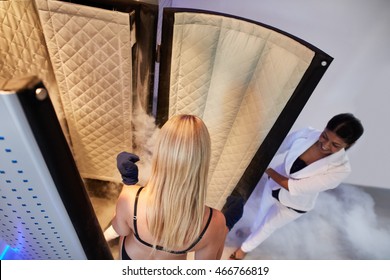 Top view of woman entering cryosauna for whole body cryotherapy. She is in lingerie, with female beautician at the cabinet door.