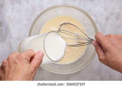 Top view of woman hand holding glass of milk putting into mixed raw eggs in glass bowl on marble surface