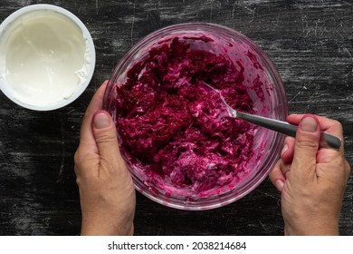 Top View Of Woman Hand Mixing Grated Cooked Beet In Glass Bowl And Sour Cream With Spoon Making Salad On Black Background