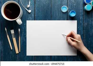 Top View Of Woman Hand With Painting Brush, Watercolor Paints And Blank Sheet Of Paper, Black Coffee Cup, Headphones. Blue Wooden Background.