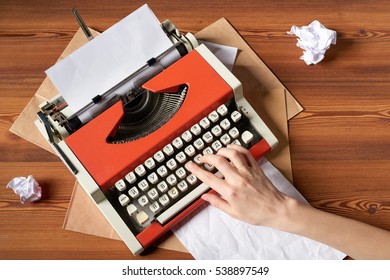 Top view of woman hand typing on red vintage typewriter with white blank paper sheet on wooden table