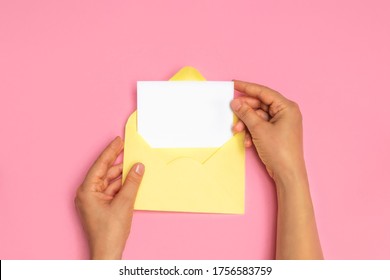 Top View Of Woman Hands Holding Empty White Card And Open Yellow Envelope Letter On Pink Background, Mockup