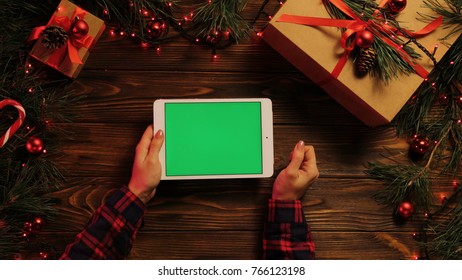 Top View Of Woman Hands Tapping, Scrolling And Zooming On Tablet Divice Horizontally. The Christmas Decorated Brown Desk With Christmas Lights. Green Screen, Chroma Key