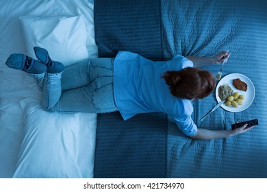 Top View Of A Woman Laying On A Bed, Eating Dinner And Watching TV