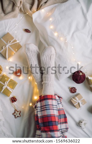 Top view of woman legs in red pajama and warm socks among decorations and gift boxes on white linen. Flat lay festive holiday concept.