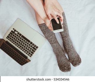 Top View Of Woman Legs In Socks And Using Smartphone With Laptop On Cozy Bed