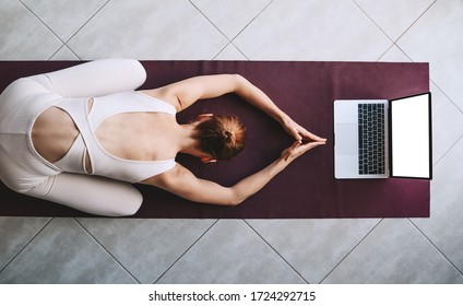 Top view of woman practicing yoga on yoga mat with laptop. Girl is meditating and relaxing with video training at home. Concept of online sport class or meditation course on digital devices.