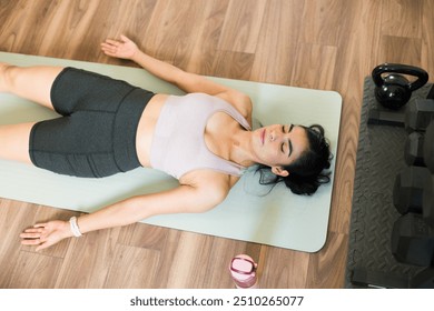 Top view of a woman is taking a break from her workout, lying on a yoga mat on the floor of her home gym - Powered by Shutterstock