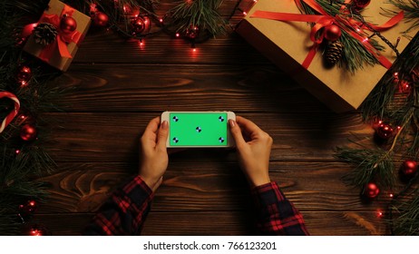 Top View. Woman Tapping, Scrolling And Zooming On The Mobile Phone Horizontally. The Decorated Wooden Desk With Christmas Lights. Green Screen, Chroma Key. Tracking Motion.