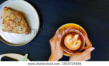 Top view of woman's hand holding art latte coffee with almond cream croissant, knife, fork and phone on dark blue wooden background or table with copy space. Flat lay of hot cappuccino drinking.