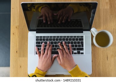 Top view of a woman's hands typing on a laptop computer, on a wooden countertop as background and a mug on the side with tea in it.