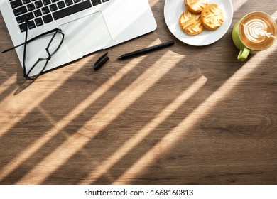 Top View Of Wood Office Desk Table With Cup Of Latte Coffee, Dish Of Almond Cookies And Office Supplies With Sun Light. Life At Work In The Morning Concept.