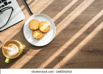 Top View Of Wood Office Desk Table With Cup Of Latte Coffee, Dish Of Almond Cookies And Office Supplies With Sun Light. Life At Work In The Morning Concept.