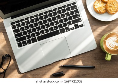 Top View Of Wood Office Desk Table With Cup Of Latte Coffee, Dish Of Almond Cookies And Office Supplies With Sun Light. Life At Work In The Morning Concept.