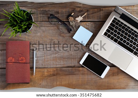 Top view of wooden desk at a workplace with a laptop computer, mobile phone, glasses, keys, post it, plant and notebook