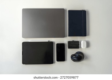 Top view workspace composition with digital devices and accessories: laptop computer, tablet, mobile phone, notebook, wallet, and wireless audio gadgets.