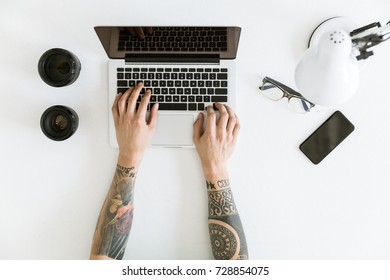 Top view of Workspace with male hands typing on laptop placed on white tabletop with camera lenses, glasses and smartphone