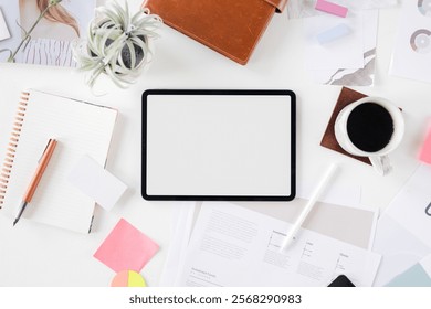 Top view of a workspace with a tablet, coffee, and stationery. Includes a blank tablet, coffee cup, and various office supplies on a white desk. Blank tablet on aesthetic workspace desk.