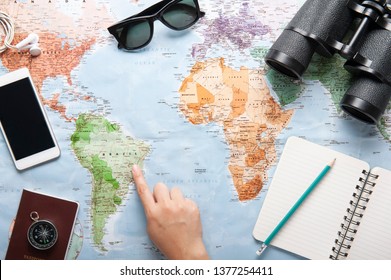 Top View Of World Map, Sunglasses, Binocular And Passport With Texting Space, Travelling Concept, Plan A Trip, Itinerary Planner, One Hand Point To The Map.