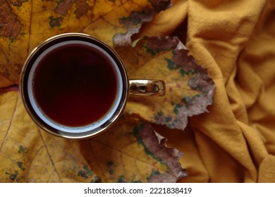 Top view of a yellow cup of tea that sits on top of a huge autumn yellow dry leaf, on a earthly tone yeallow cotton scarf