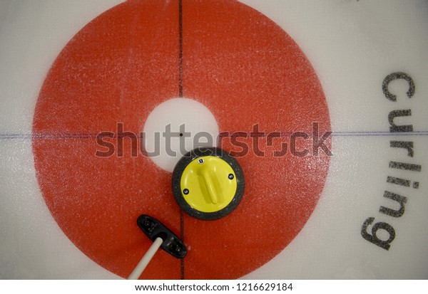 Top view of yellow curling\
stone in inner red ring of the house, near the button, with broom\
nearby.