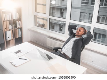 Top View Of Young African Guy Is Sitting At Office In Chair And Reclining On The Back. He Smiling While With Relaxation Looking At Ceiling. Copy Space In Left Side