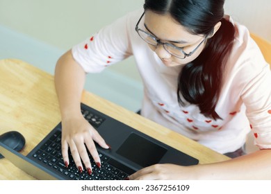 Top view of young Asian female with glasses writer keyboarding on laptop while working for online writing work. People, lifestyle, modern technology and communication concept.