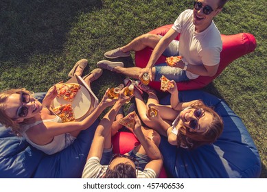 Top View Of Young Beautiful People In Casual Clothes And Sun Glasses Eating Pizza, Drinking, Looking At Camera And Smiling, Sitting On Bean Bag Chairs While Resting Outdoors