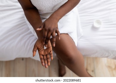 Top view of young black lady applying hand cream, sitting on bed after bath, wearing towel, pampering herself at home. Overhead shot of Afro woman making skin-care treatment