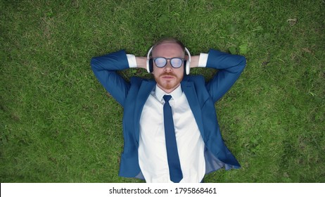 Top view of Young Businessman Lying On Grass Enjoying Music On Headphones In Park. Attractive guy in formal suit listening to music in earphones relaxing on green lawn