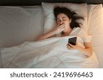 Top view of young Caucasian woman using smartphone and yawning, lying in bed at night. Insomnia, sleepless and social media addiction.