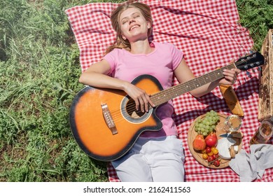 Top View Of Young Caucasian Woman In White Pants Outside Having Picnic, Eating And Playing Guitar. Summer Fun And Leisure