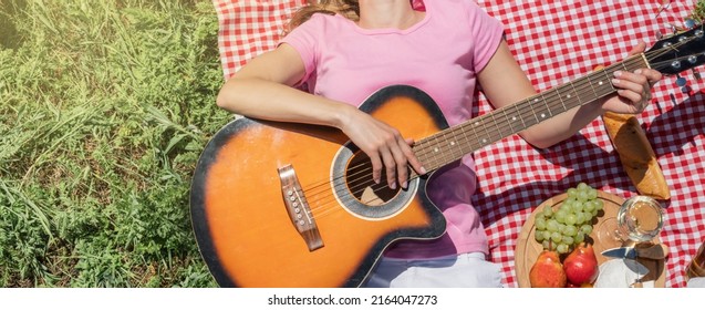 Top View Of Young Caucasian Woman In White Pants Outside Having Picnic, Eating And Playing Guitar. Summer Fun And Leisure