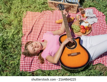 Top View Of Young Caucasian Woman In White Pants Outside Having Picnic, Eating And Playing Guitar. Summer Fun And Leisure