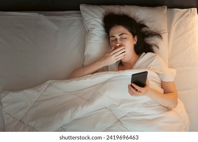 Top view of young Caucasian woman using smartphone and yawning, lying in bed at night. Insomnia, sleepless and social media addiction. - Powered by Shutterstock