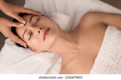 Top view of young female with bare shoulders lying on bed while her head is being massaged by professional face masseur at spa