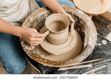Top view of young female potter in apron making shape of clay vase with wooden tool on spinning pottery tool in ceramic workshop, clay shaping and forming process