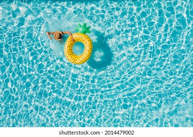 Top View Of A Young Female In Swimsuit Bikini In A Straw Hat On Blue Swimming Pool Waves Background With Big Inflatable Yellow Pineapple Tube. Chill Out A Summer Vacation In Luxury Resorts Concept.