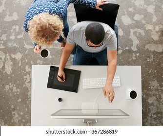 Top view of young graphic designers working in office. Using digitized graphic tablet, digitized pen and desktop computer. Man showing his work on monitor to woman standing by with a cup of coffee. - Powered by Shutterstock