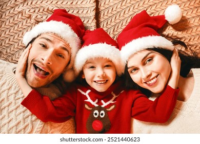 Top view of young happy family in santa hats laying down on bed or floor, little girl embracing parents, happy family concept - Powered by Shutterstock