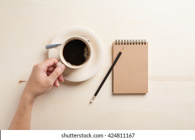 Top view of Young hipster male left hand holding coffee cup on wood table beside small notebook with blank area on brown cover in morning time - Powered by Shutterstock